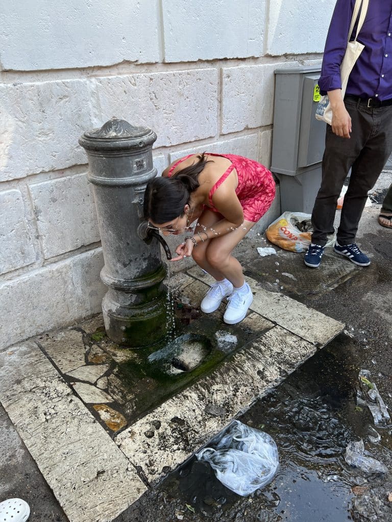 Woman in red dress bending to drink from a nasone or public drinking foutain in Trastevere.