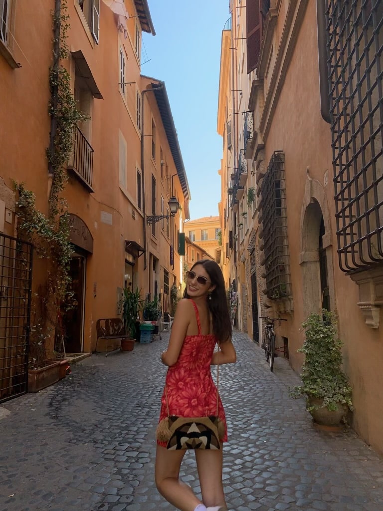 Girl in red dress looking over her shoulder walking down a narrow cobblestone street in Rome.