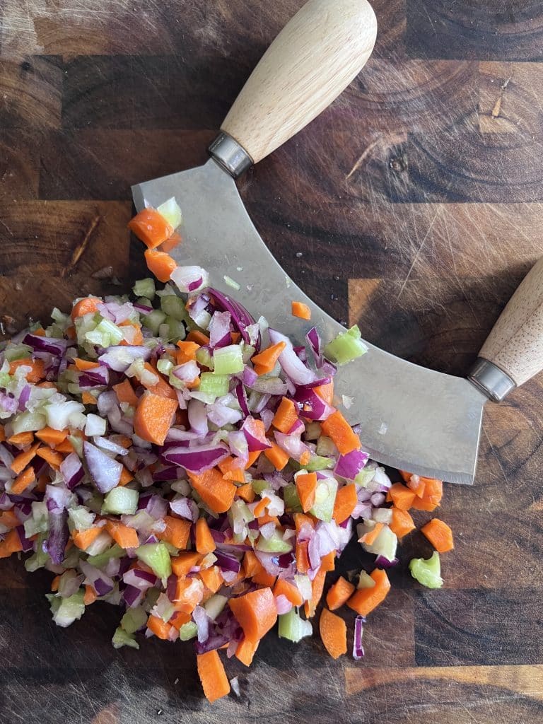 Finely chopping celery, carrot, end red onion using a mezzaluna chopping tool on a cutting board.