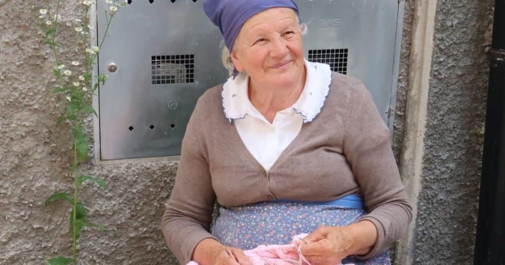An elderly woman knitting outside her home on a street in Schilpario, Italy