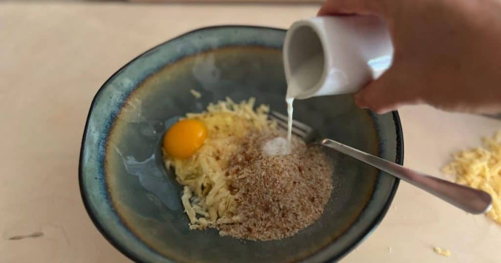 Hand pouring milk on top of breadcrumbs, cheese, and egg.